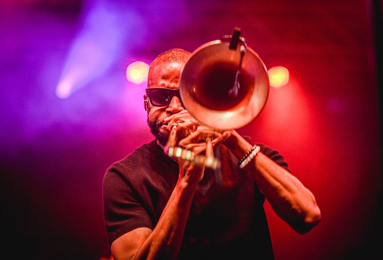 Trombone player in sunglasses with the bell pointed toward the camera under purple and red stage lights